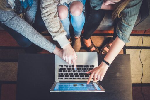 Ein Foto von 3 Personen, von denen man nur die Beine sieht. Sie zeigen mit ihren Händen auf einen Laptop.