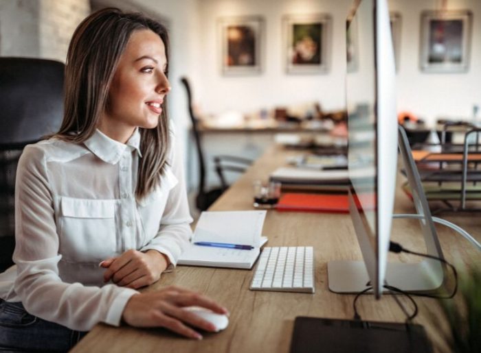 A photo of a woman who looks at her computer screen and smiles.