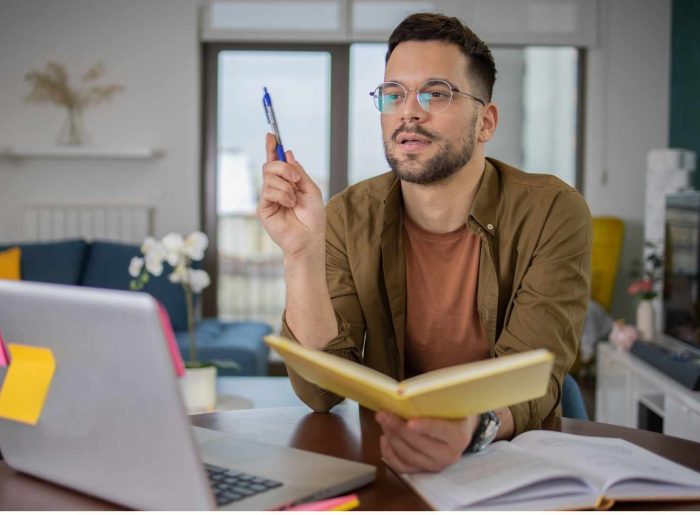 A man with glasses and a brown shirt is sitting in front of a laptop with a book in his hand.