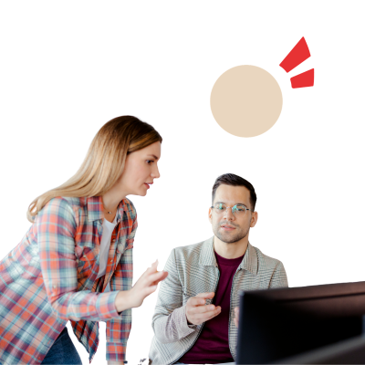A photo of two people. One person is sitting in front of the screen, and the other is standing next to it. They are talking to each other.